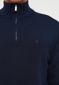 Mann iført en marineblå genser med kvart glidelås og et lite brodert "cK"-logo på brystet.