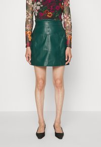 Grön läder mini kjol med hög midja, plisserade detaljer, tryckknappsstängning och två framfickor. Slät textur.