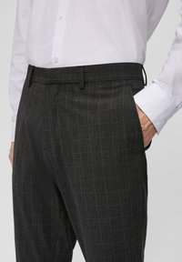 Pantalons gris à carreaux en tissu texturé, présentant une taille plate, des poches latérales et une coupe cintrée soulignée par des lignes subtiles.