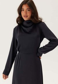 Abito a maniche lunghe e collo alto in morbido tessuto nero, con vita aderente e silhouette elegante. Texture morbida con dettagli minimi.