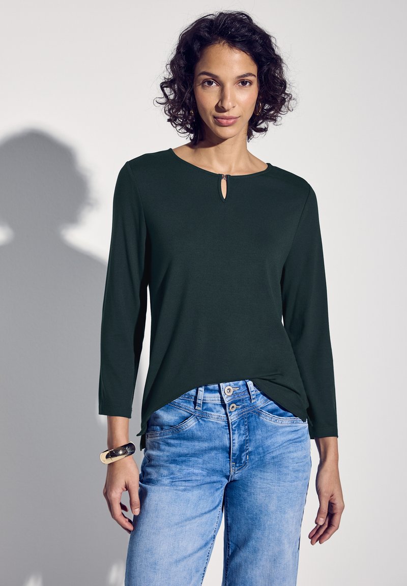 Dunkelgrünes Langarmtop mit einem Schlüsselaugen-Detail am Ausschnitt, kombiniert mit hellblauen Jeans. Sanfte Textur, lässige Passform.