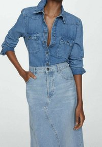 Camicia di denim di blu medio con bottone frontale e due tasche sul petto. Abbinata a una gonna di denim blu chiaro coordinata, entrambe con un taglio classico.