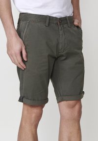 Vīrietis, kuram ir uzreiz pārstrādātas olīvzaļas šortus ar sānu kabatām, kombinēts ar baltu t-kreklu, rokas tuvumā kabatām.