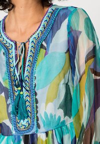 Barevná blůza s květinovým vzorem. Má výstřih do V, dekorativní šití a třásně v tyrkysové a zelené barvě. Průhledná, lehká látka.
