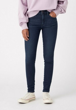 Osoba ubrana w ciemnoniebieskie obcisłe dżinsy, jasnofioletową bluzę i białe wysokie trampki, stojąca z jedną ręką w kieszeni.