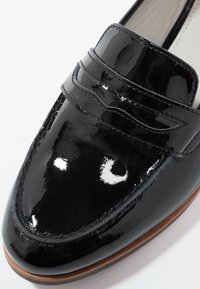 Mocassin en cuir verni noir avec bride à penny, bout arrondi et bord de semelle marron, présentant une finition brillante et des coutures détaillées.