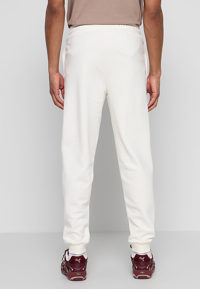 Pantalones deportivos blancos con cintura elástica, piernas entalladas y puños acanalados. Se aprecia la textura suave de la tela. Usados con zapatillas deportivas burdeos.