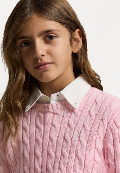 Ανοιχτό ροζ πουλόβερ με πλέξη καλωδίου και στρογγυλή λαιμόκοψη, επικαλυμμένο πάνω από ένα λευκό πουκάμισο με γιακά. Σημειώστε την εμφανή ριμπ ύφανση και την λεπτομέρεια του λογότυπου στο στήθος.