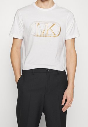 Homme portant un t-shirt blanc à col rond avec un logo "MK" doré et un pantalon noir taille haute, mains dans les poches, sur un fond uni.