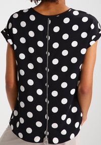 Svart blus med vita prickar, korta ärmar och dragkedja i ryggen. Slät tyg med en avslappnad passform.