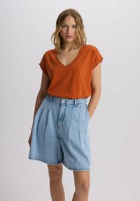 Rdzawopomarańczowa koszulka V-neck z krótkimi rękawami, w połączeniu z jasnoniebieskimi spodniami dżinsowymi o wysokim stanie i plisami. Gładki materiał i luźny krój.