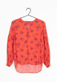 Blouse rouge à motif floral, avec des manches longues, un dos en clé et un ourlet inférieur transparent. Fabriquée en tissu léger et texturé.