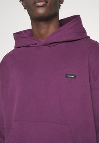 Paarse katoenen hoodie met een trekkoordcapuchon, een voorzak en een zwart rechthoekig logo-opdruk op de borst. Gladde textuur.