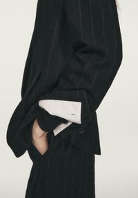 Czarna marynarka w prążki o lekko oversize'owym kroju, z białym mankietem koszuli wystającym spod podwiniętego rękawa oraz teksturowanymi detalami materiału.