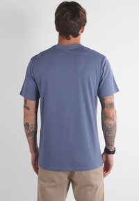 Homme avec des cheveux bruns portant un t-shirt bleu et un pantalon beige, montrant des bras tatoués et une montre noire au poignet, se tenant de dos.