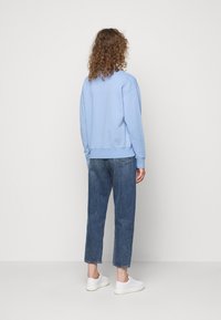 Ljusblå sweatshirt med rund halsringning, ribbade ärmslut och nederkant, i kombination med mörkblå avsmalnande jeans och vita sneakers. Lockigt hår synligt.