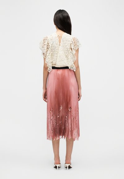 Haut en dentelle avec manches à volants couleur crème, associé à une jupe plissée rose ornée d'accents floraux. Ceinture noire et talons blancs.