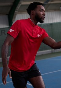 Polo corto rosso a maniche corte con colletto e dettagli a bottoni, con un logo sul petto. Indossato con pantaloni corti neri, su un campo da tennis.