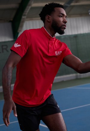 Red short-sleeve polo with a collar and button detail, featuring a logo on the chest. Worn with black shorts, on a tennis court.