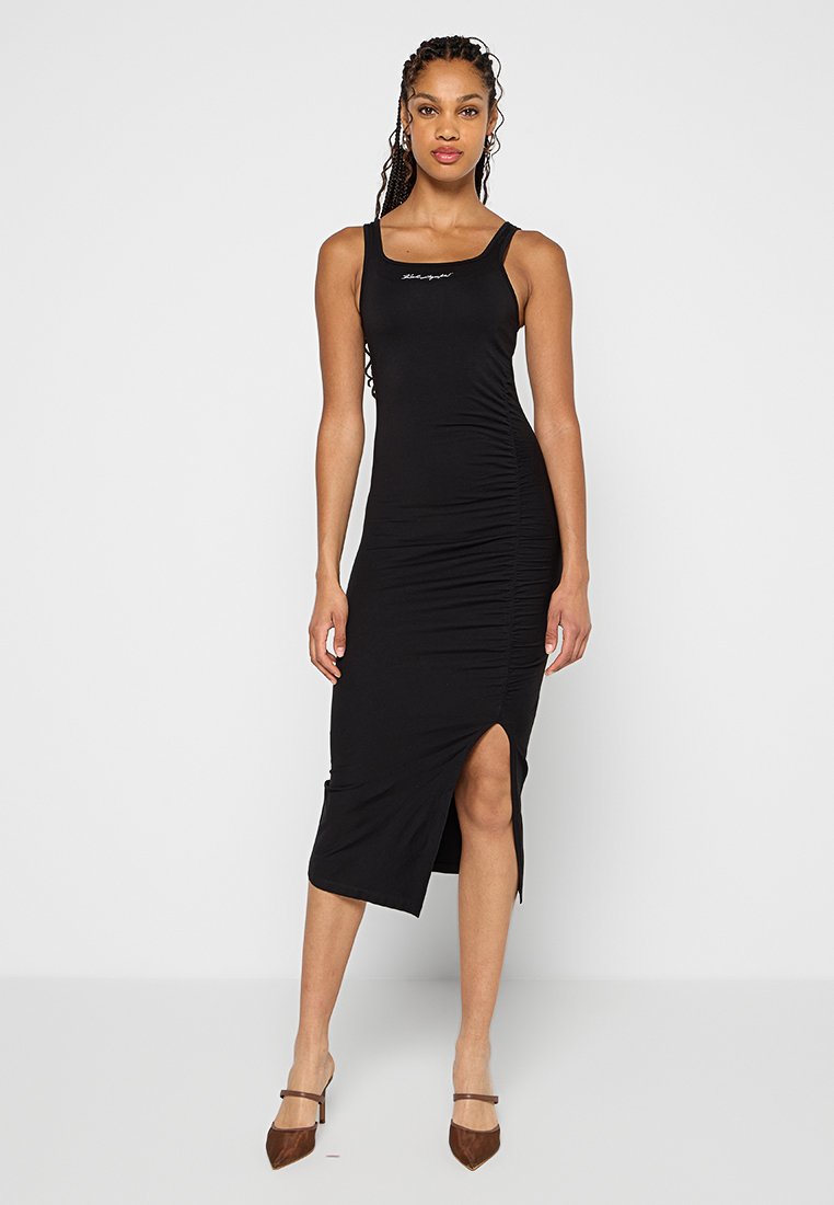 Frau in einem ärmellosen schwarzen Midikleid mit seitlicher Raffung und Schlitzen bis zum Oberschenkel, die braune spitz zulaufende Absätze trägt und vor einem weißen Hintergrund steht.