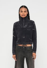 Īsāks, tumši zils džemperis ar augstu apkakli un puszipu, kas izceļas ar teksturētu dizainu. Apakšmalā ir aukliņa ērtākai pielāgošanai.