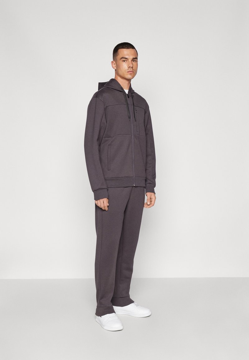 Conjunto deportivo de dos piezas para hombres en gris oscuro. La chaqueta presenta un cierre de cremallera frontal, capucha y bolsillos frontales. Los pantalones son ajustados con puños acanalados. Zapatillas blancas.