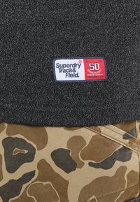 Mørkegrå stof med en hvid og rød Superdry Track & Field-label over tan- og brunt camouflage-stof med synlige sømme.