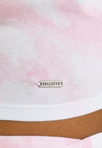 Camisa de algodón con un patrón de tie-dye en rosa y blanco, con una etiqueta metálica marcada "Hollister" cerca del dobladillo. Tela suave y ligera.