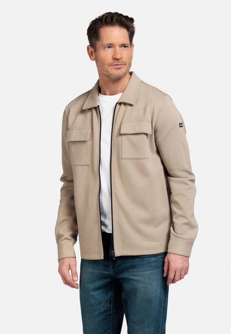 Beige zip-up jack met zachte stof, voorzien van twee voorzakken, geribbelde boorden en een staande kraag, gecombineerd met een wit T-shirt en blauwe spijkerbroek.
