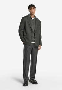 Grau strukturierte Blazer mit zwei Knöpfen, über einem grauen Hoodie getragen. Kombiniert mit grauen Hosen und schwarzen Schuhen. Sauberer, moderner Silhouette.