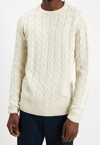 Maglione a trecce color crema, con scollatura e polsini a coste. Realizzato in tessuto testurizzato con una vestibilità aderente e un piccolo logo decorativo.