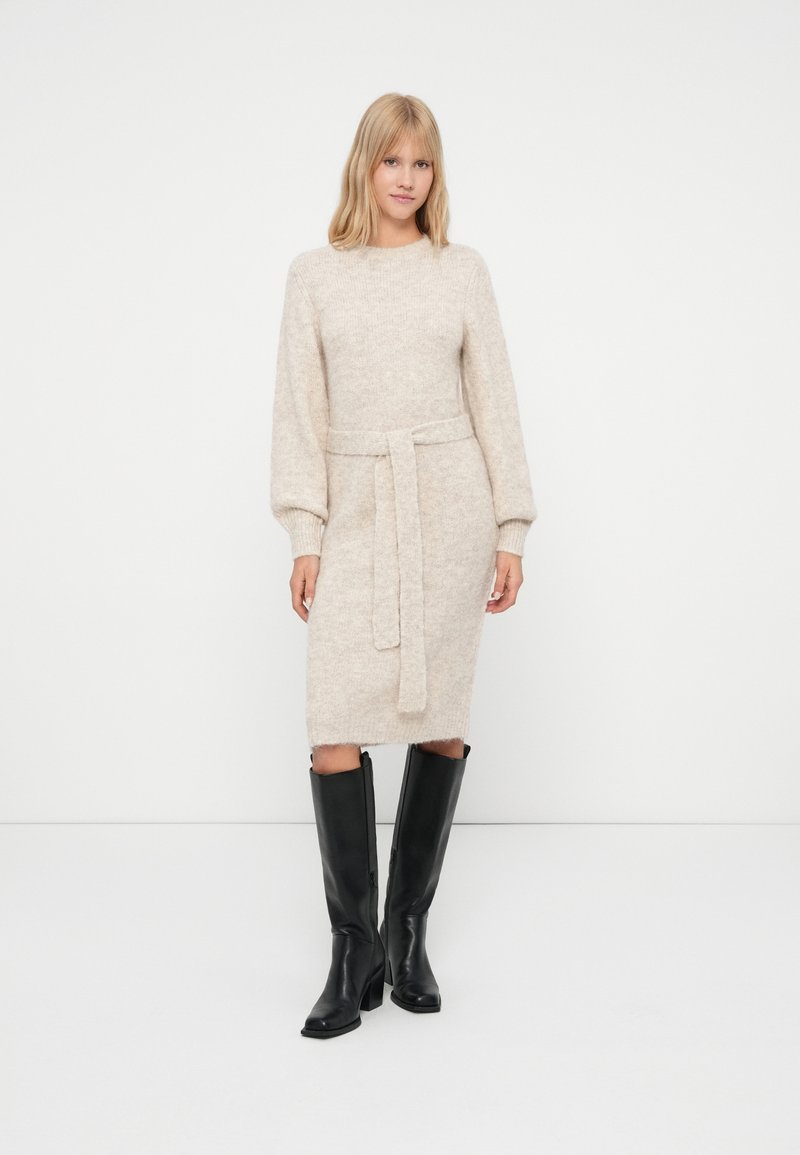 Abito maglione beige lavorato a maglia con maniche a sbuffo lunghe e vita cintrata. Presenta una linea dritta e polsini a costine, abbinato a stivali neri sopra il ginocchio.