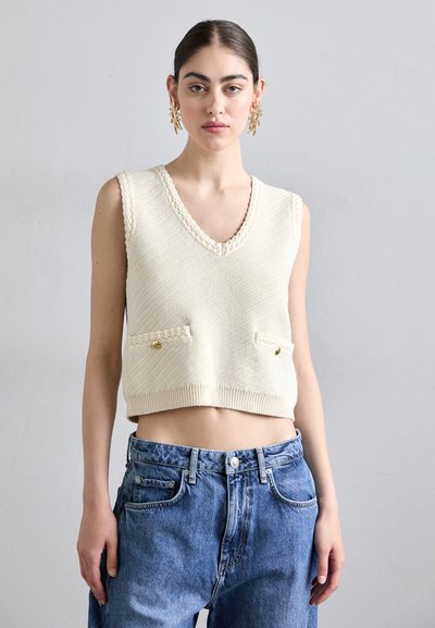 Débardeur crème en tricot sans manches avec un col en V, un motif texturé et deux poches avant ornées d'accents dorés, associé à un jean en denim taille haute.