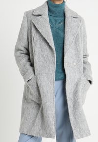 Cappotto in misto lana grigio con rever a punta, chiusura doppiopetto, tasche laterali e una vestibilità rilassata, abbinato a un dolcevita texturizzato color teal.