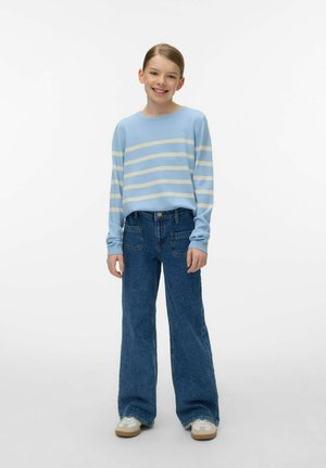 Młoda dziewczyna stojąca, uśmiechnięta, ubrana w jasnoniebieski sweter w białe paski, szerokie niebieskie dżinsy i białe trampki na jednolitym tle.