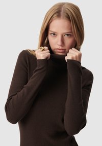 Maglione a collo alto marrone a costine con maniche lunghe. Modello che tiene il colletto, mettendo in mostra la texture e la vestibilità. Anello d'oro sulla mano sinistra.