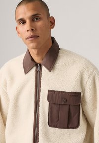 Chaqueta de forro polar color crema con un cuello marrón y un bolsillo frontal con solapa. Cierre de cremallera y detalle de tejido texturizado por toda la prenda.