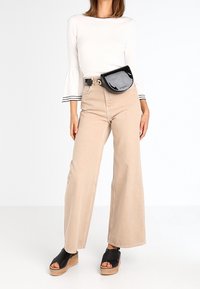 Bílá košile s dlouhým rukávem a zvonovými manžetami, béžové široké kalhoty, černá ledvinka a černé platformové sandály se dřevěnými podrážkami.