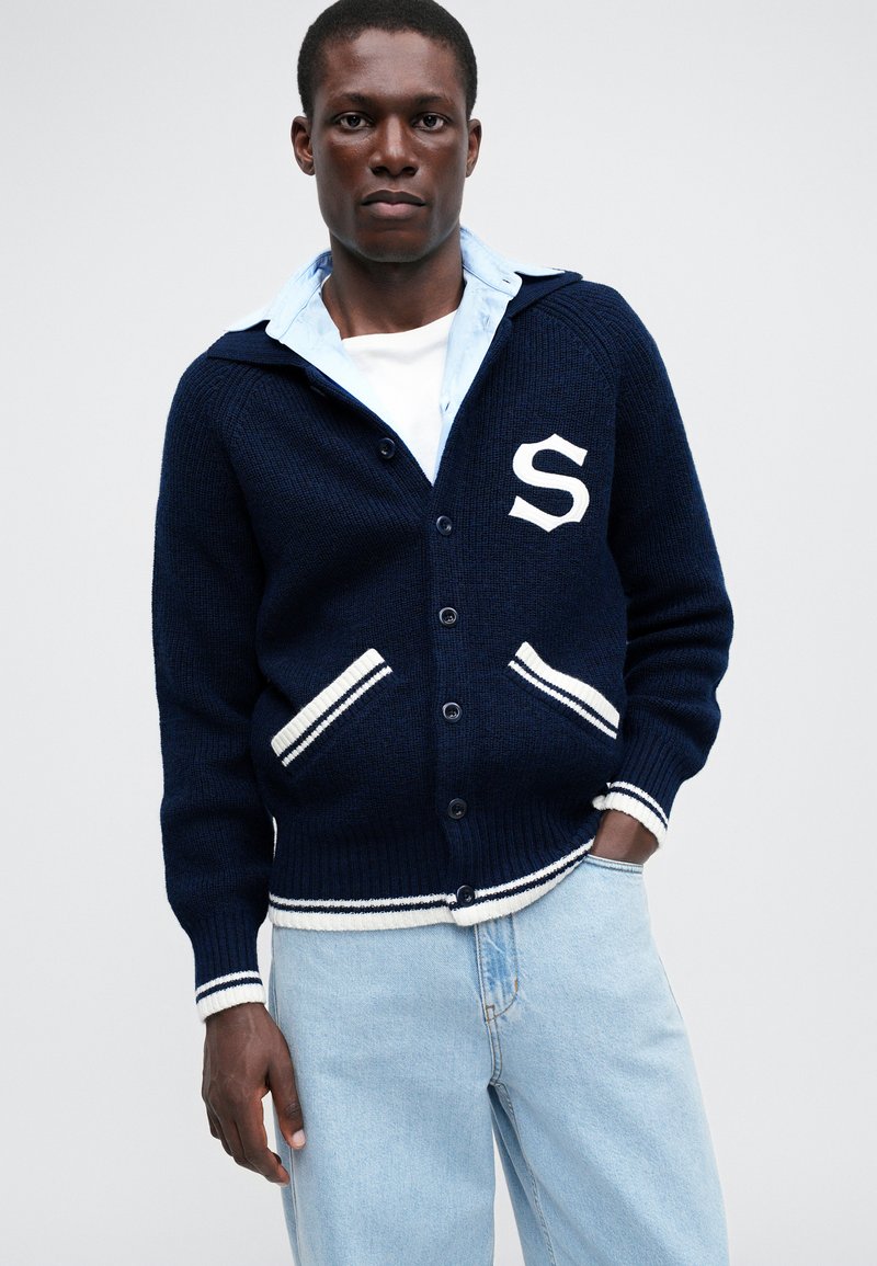 Marine gebreide cardigan met witte strepen, voorzien van een capuchon, voorzakken en een wit "S" embleem. Gecombineerd met een lichtblauw shirt en denim.