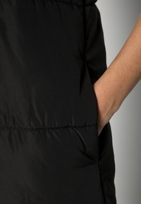 Czarna pikowana kamizelka o gładkiej, błyszczącej fakturze; posiada boczną kieszeń z widocznym przeszyciem i dopasowany fason.