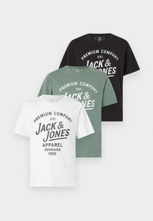 Muž v bielom tričku s logom Jack & Jones, béžových cargo nohaviciach, čiernych teniskách s bielymi pruhmi a čiernych slnečných okuliaroch stojící na bielom pozadí.