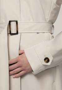 Beige trenchcoat met een riem met een rechthoekige, donkere gesp. De manchet heeft een ronde, donkere knop en gedetailleerde stiksels.
