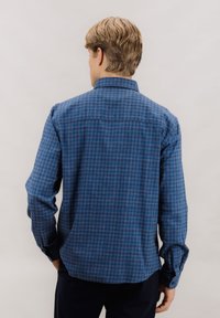 Blå rutete skjorte med knapp-down krage, lange ermer og rundet nederkant. Stoffet har en teksturert overflate med varierende nyanser av blått.