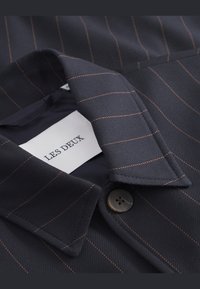 Cămașă customizată bleumarin cu dungi portocalii, având un guler clasic și o închidere cu un singur nasture aproape de guler. Eticheta mărcii este vizibilă.
