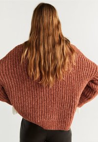 Pull-over surdimensionné et texturé dans une couleur rouille chaude. Présente des manches larges et une coupe ample, avec un détail côtelé sur tout le tissu.