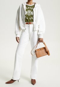 Sudadera blanca con cremallera, camiseta gráfica verde oliva, vaqueros blancos de talle alto, bolso de cuero marrón y tacones marrones puntiagudos con un diseño texturizado.