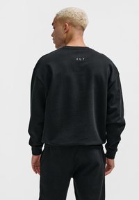Svart sweatshirt med nedhasade axlar, rund halsringning och broderad text "F.U.T." på baksidan. Materialet verkar mjukt med en matt finish.