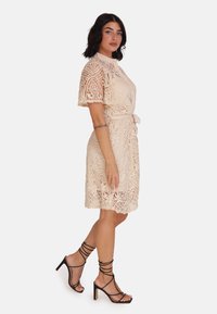 Sukienka koronkowa w jasnym beżu z krótkimi rękawami, paskiem w talii i misternymi wzorami. Połączona z czarnymi sandałami na obcasie typu block-heel z paskami.