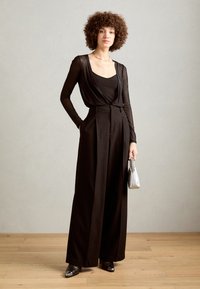 Black long-sleeve top with sheer fabric layered over a fitted black tank, paired with high-waisted black wide-leg trousers and silver handbag.