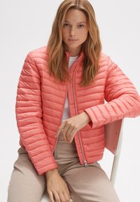Veste en corail clair, gonflante, avec texture matelassée, fermeture éclair intégrale et col montant. Associée à un pantalon beige dans un cadre minimaliste.